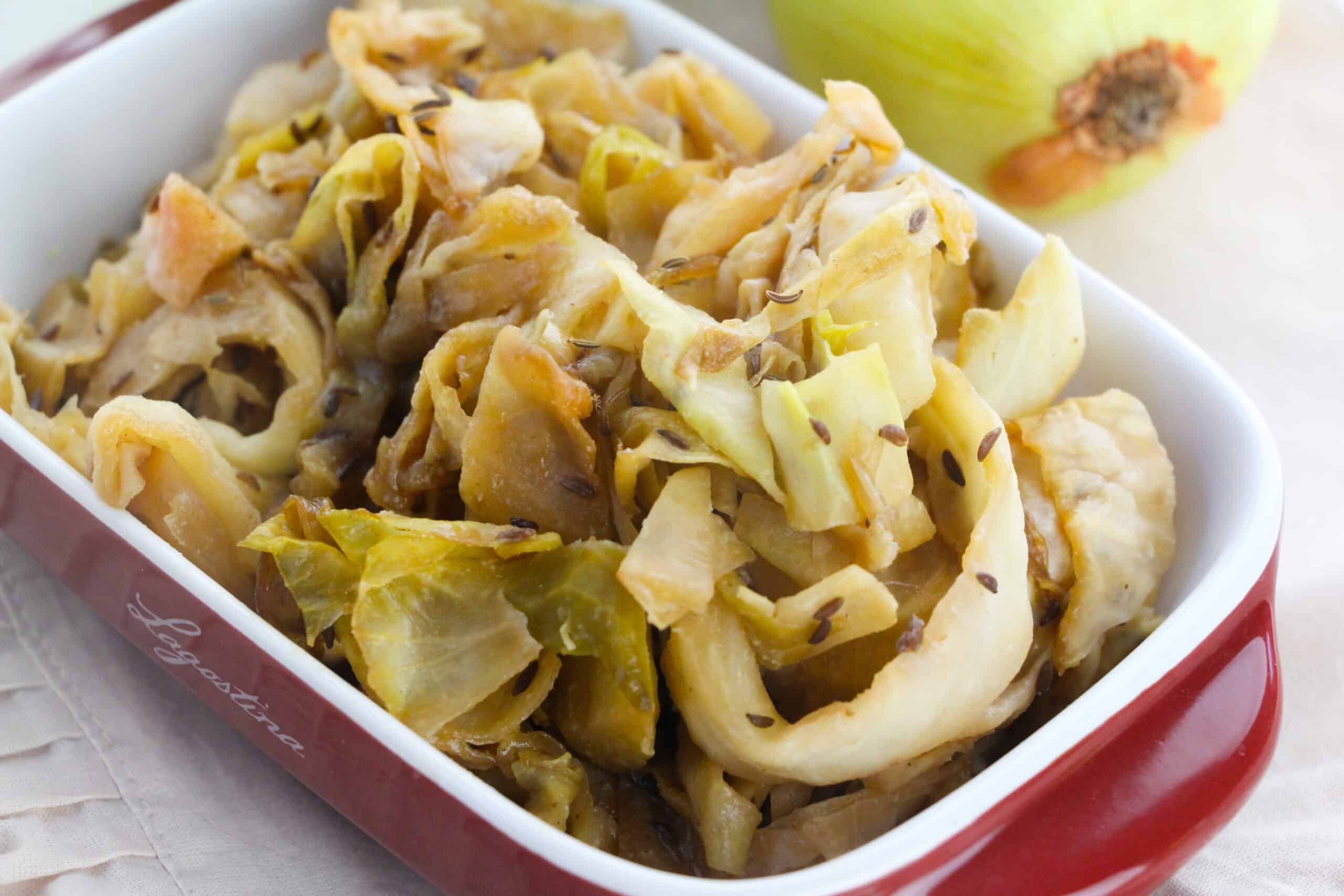 Braised cabbage with caraway seeds is served in a red and white ceramic dish, with an onion in the background.