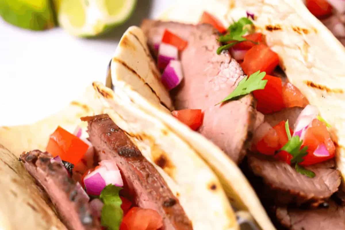 Three steak tacos topped with diced tomatoes, red onions, and cilantro are arranged closely together on tortillas. Packed with flavor, this is a must try for anyone who loves authentic street food recipes. Lime wedges are visible in the background.