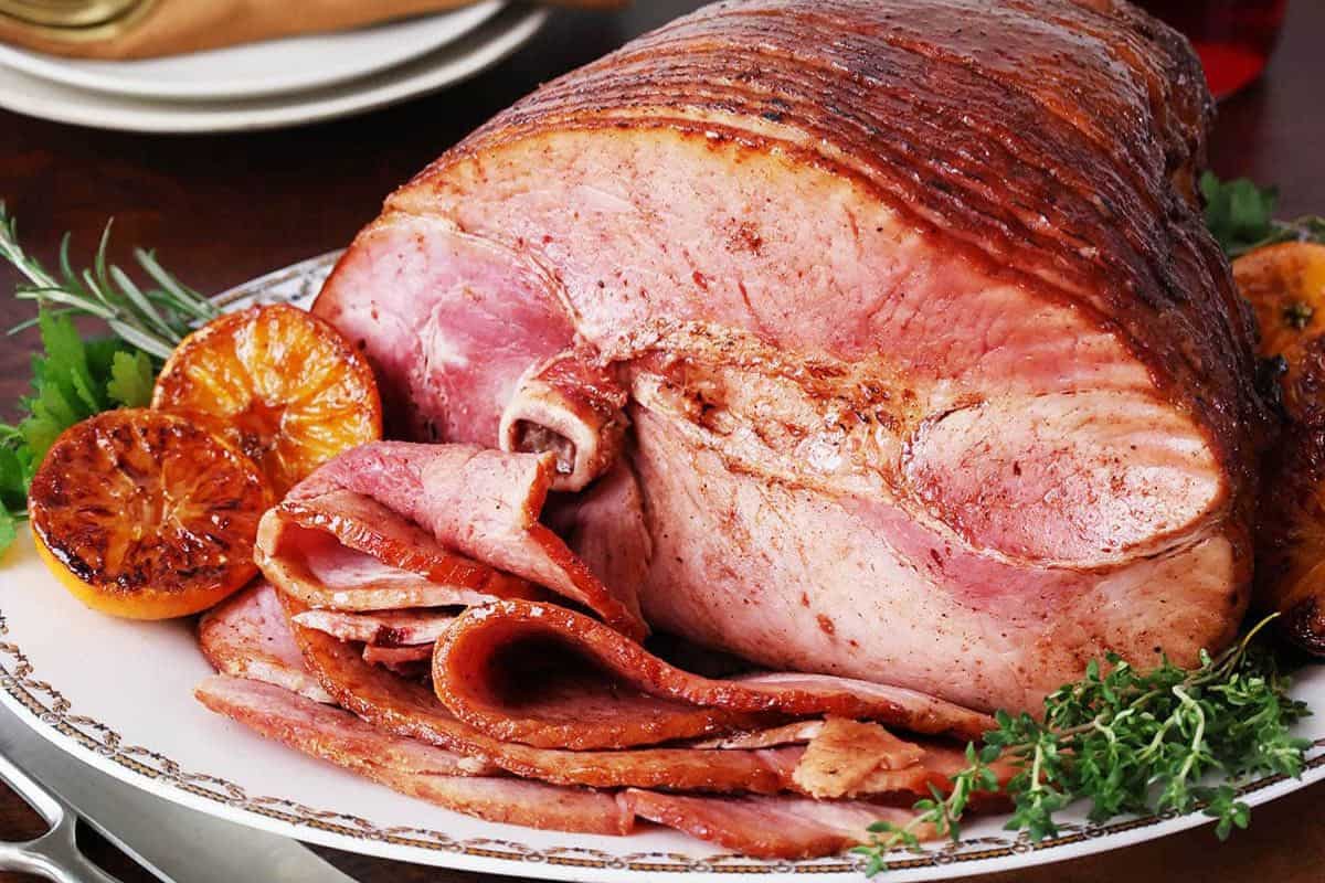 A large glazed ham is sliced and served on a platter with herbs and roasted orange slices—perfect for any gathering and a standout among Holiday Ham Recipes.