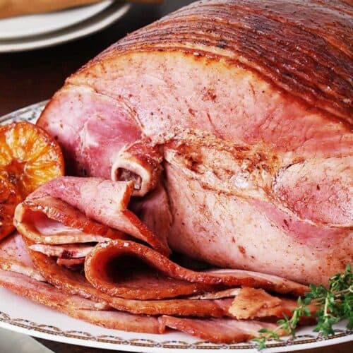 A sliced glazed ham is served on a platter with herbs and roasted orange halves, making it a standout among classic holiday ham recipes.