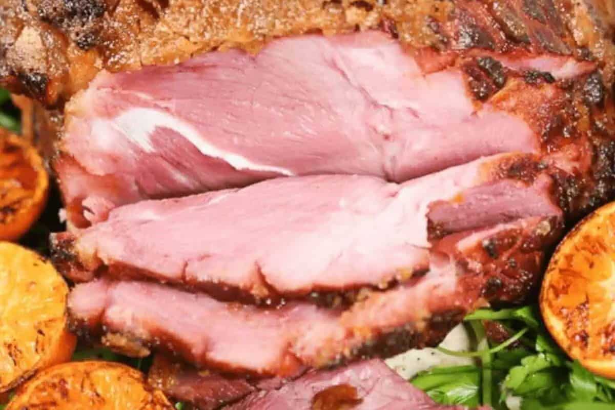 Close-up of sliced, cooked ham with a browned crust, garnished with grilled orange slices and fresh herbs—a perfect centerpiece for holiday ham recipes.