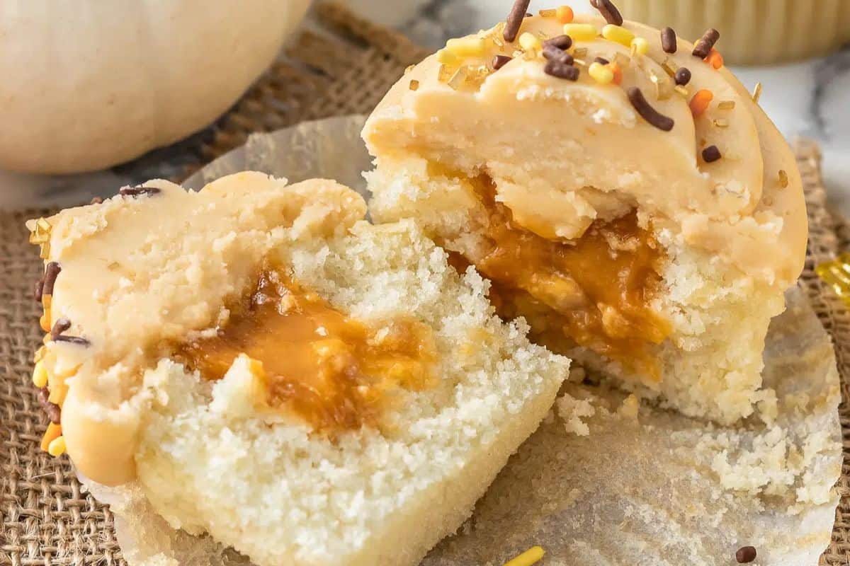 Cupcake cut in half, revealing a deliciously rich caramel filling. Topped with beige frosting and sprinkled with brown, yellow, and orange sprinkles. Perfect for butterscotch recipe lovers—try today on a textured surface.