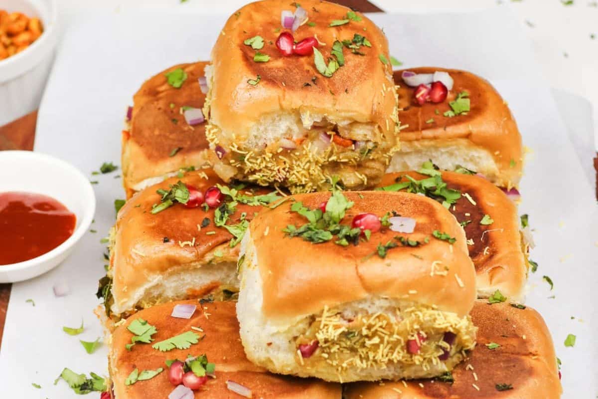 A stack of vada pav, a must try from street food recipes, garnished with sev, chopped onions, cilantro, and pomegranate seeds, served with a side of red chutney for an extra burst of flavor.