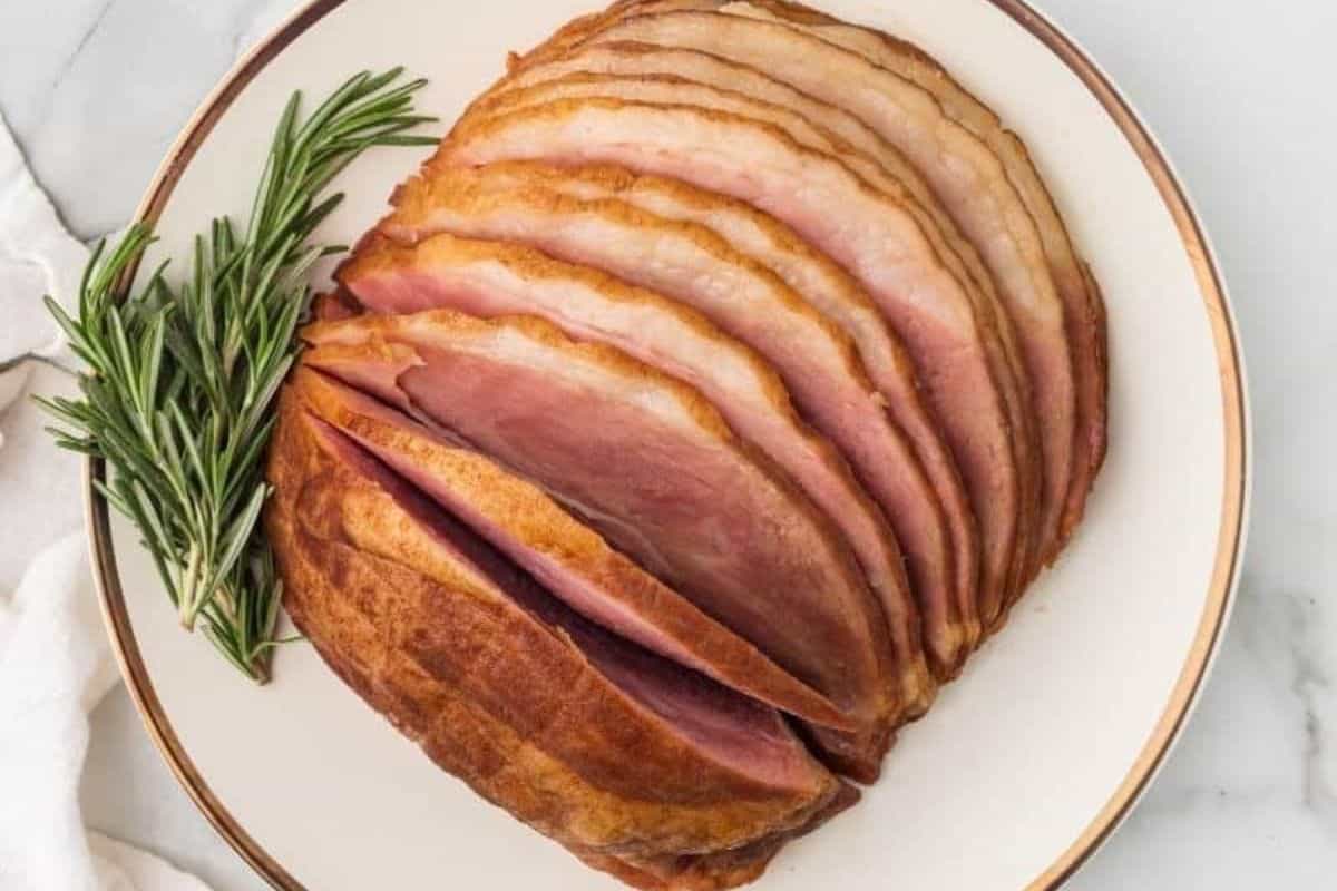 Sliced glazed ham arranged on a white plate with a sprig of fresh rosemary on the side—perfect for classic ham recipes and festive holiday gatherings.