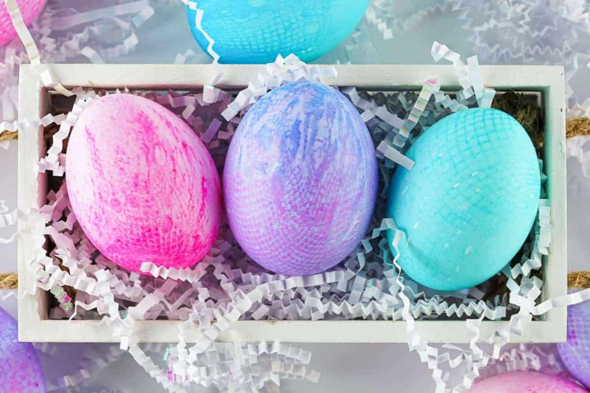 Three pastel-colored Easter eggs—pink, purple, and blue—sit in a white rectangular tray filled with white paper shreds, making for charming Easter egg decorating inspiration and easy Easter crafts.