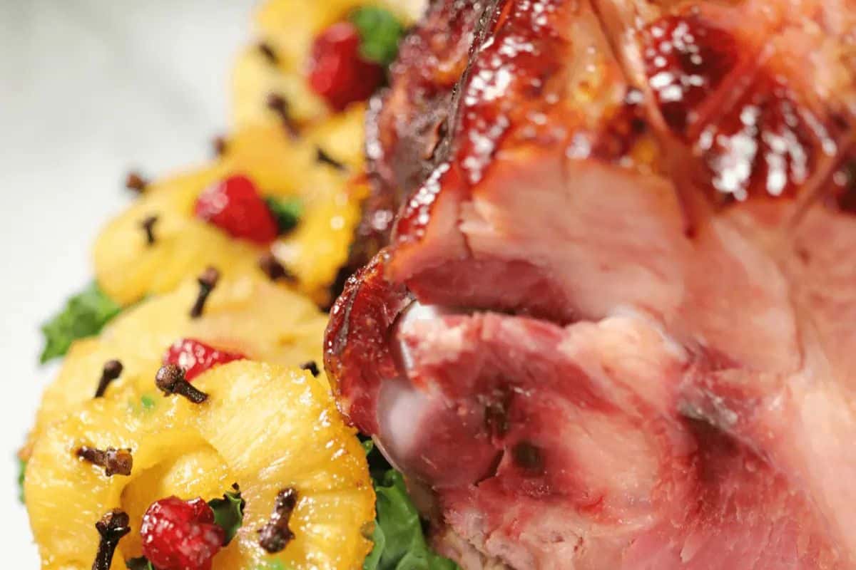 A classic holiday ham is studded with cloves, ringed with glossy pineapple slices and festive red cherries, resting on a bed of greens. The ham is deeply browned and juicy, with a hint of sweet glaze over its scored surface. This traditional presentation screams holiday nostalgia.
