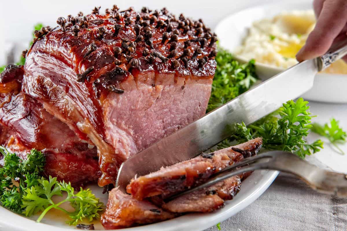 A glazed ham studded with cloves is being sliced on a white plate, garnished with fresh parsley, with mashed potatoes in the background—a delicious centerpiece perfect for classic ham recipes or your favorite holiday recipes.