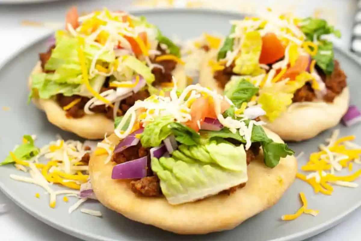 Three fry bread pieces topped with ground meat, lettuce, tomato, red onion, and shredded cheese are arranged on a gray plate—a must try street food for fans of flavorful street food recipes.