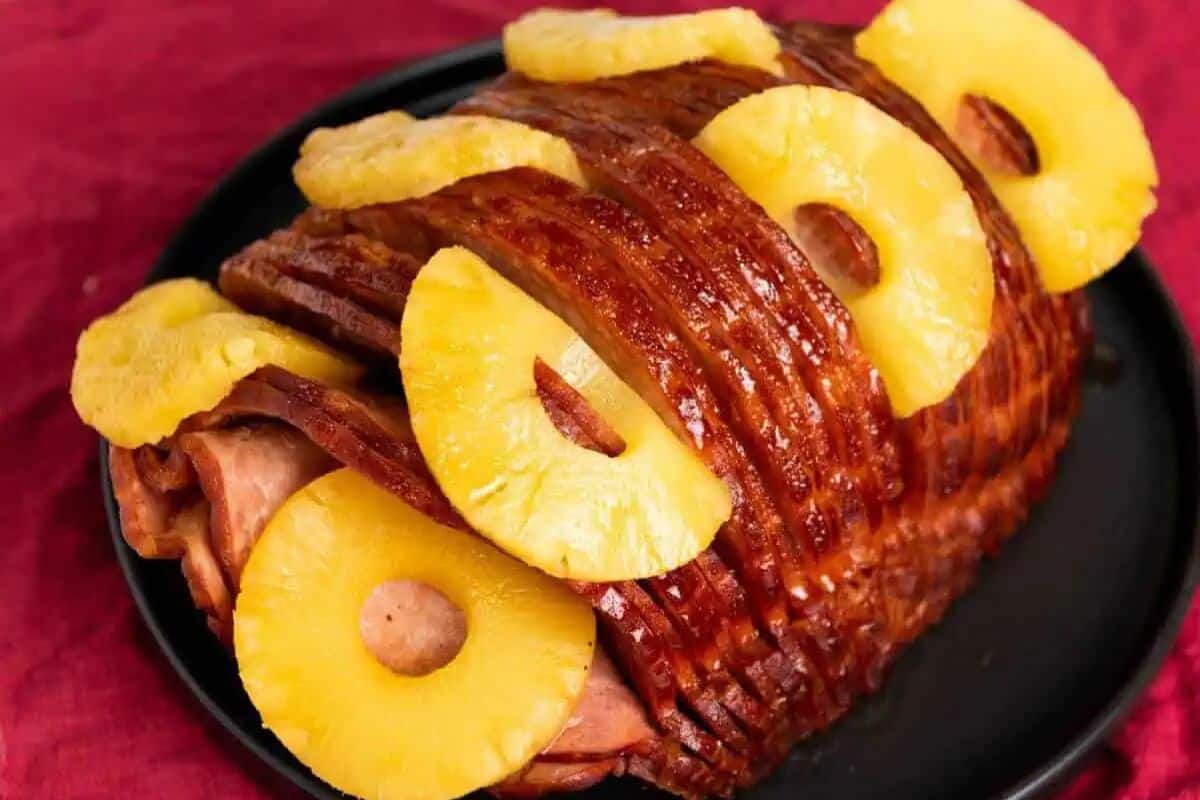 Sliced glazed ham on a black plate, topped with several round pineapple rings, set on a red surface—perfect for classic ham recipes or your favorite holiday ham recipes.