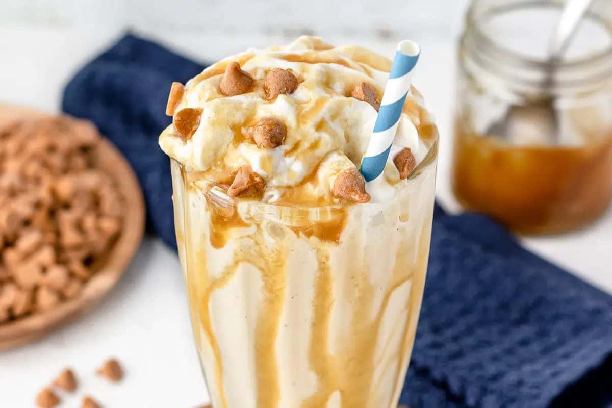 Indulge in this deliciously rich milkshake topped with whipped cream, caramel drizzle, and caramel chips, served with a striped straw. A jar of caramel and a bowl of chips temptingly await on a blue cloth. Try today for the ultimate treat!.