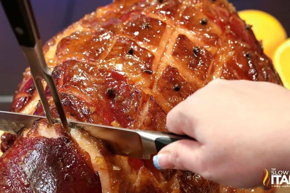 A person slices a glazed, baked ham with a knife; the ham, perfect for holiday ham recipes, features a crisscross pattern and is studded with cloves and oranges in the back.