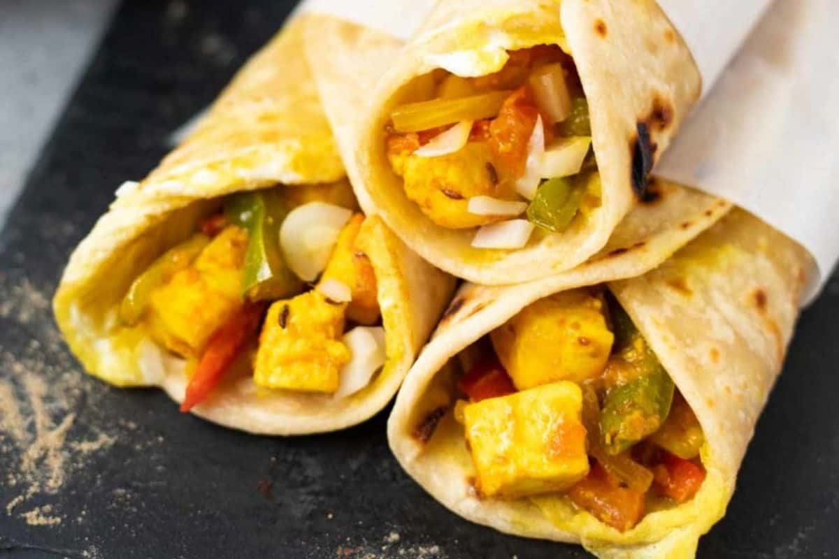 Three paneer wraps filled with flavorful paneer cubes, sliced onions, and bell peppers, wrapped in flatbread and placed on a dark surface—a must try for lovers of street food recipes.