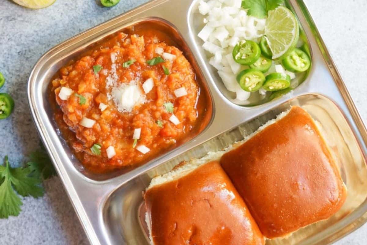 Stainless steel tray with pav bhaji, chopped onions, green chilies, lime wedge, and two buttered bread rolls—a street food recipe that’s a must try for flavorful recipes lovers.