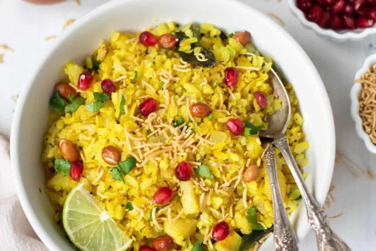 A bowl of yellow poha, a flavorful street food, is garnished with peanuts, sev, pomegranate seeds, cilantro, and a lime wedge, with two spoons placed inside.