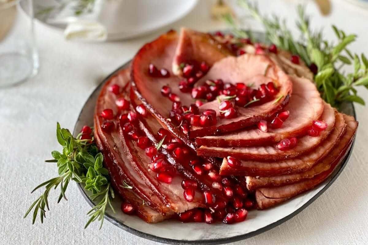 Sliced ham arranged on a plate, garnished with fresh herbs and topped with pomegranate seeds—a festive touch for your favorite holiday ham recipes.