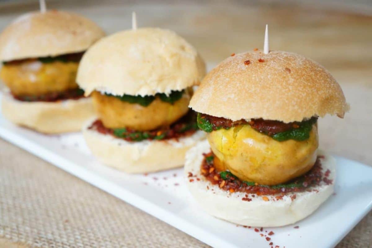 Three slider-sized burgers with round patties, green herbs, and red sauce on white buns, each secured with a toothpick—these flavorful street food bites are artfully arranged on a rectangular white plate.