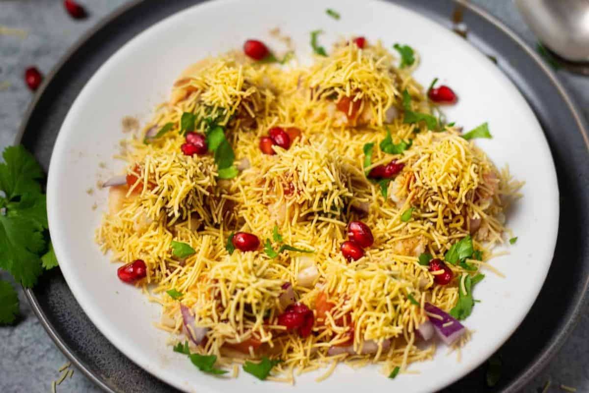 A plate of Indian chaat, a must try street food, garnished with sev, chopped onions, cilantro, and pomegranate seeds on a white dish.