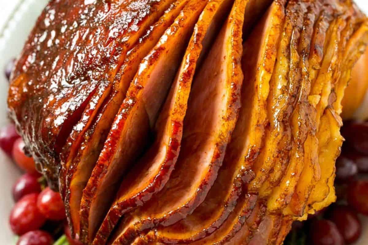Sliced glazed ham with a shiny, caramelized surface, displayed on a platter with red grapes around the base—a perfect centerpiece for Holiday Ham Recipes.