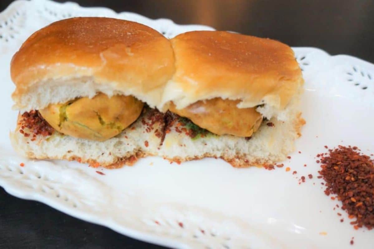 Two vada pav sandwiches on a white plate, each with a fried potato patty inside a bread bun, and some red chili powder sprinkled on the side—a classic street food recipe bursting with flavor.