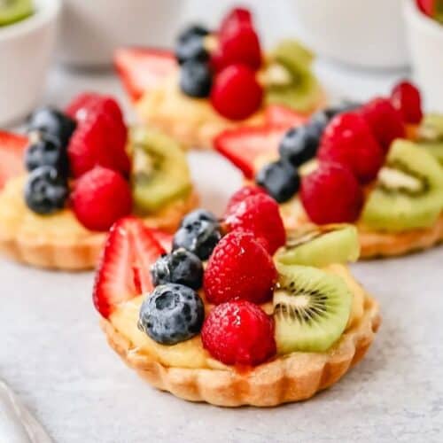 Mini fruit tarts with custard filling topped with fresh strawberries, blueberries, raspberries, and kiwi slices—an irresistible treat perfect for Summer Dessert Recipes. Arranged beautifully on a light surface.