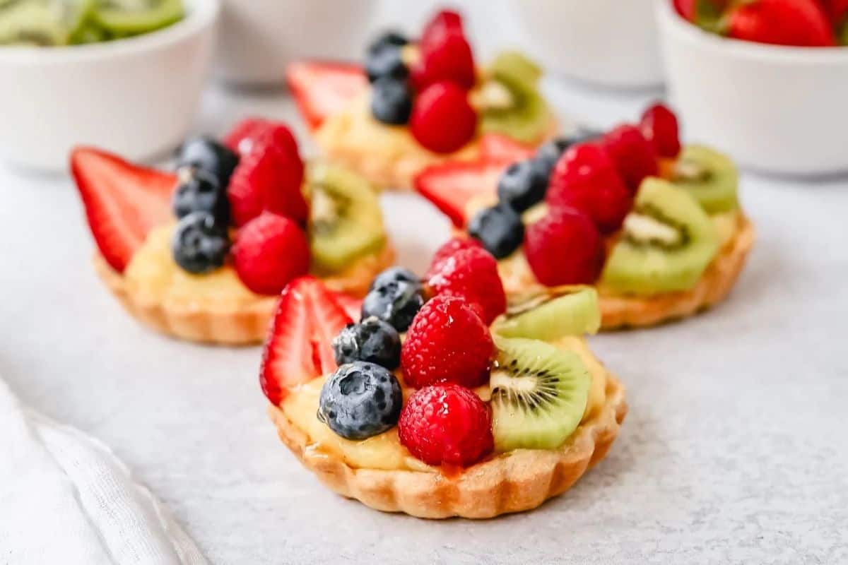 Mini fruit tarts with custard filling topped with fresh strawberries, blueberries, raspberries, and kiwi slices—an irresistible treat perfect for Summer Dessert Recipes. Arranged beautifully on a light surface.