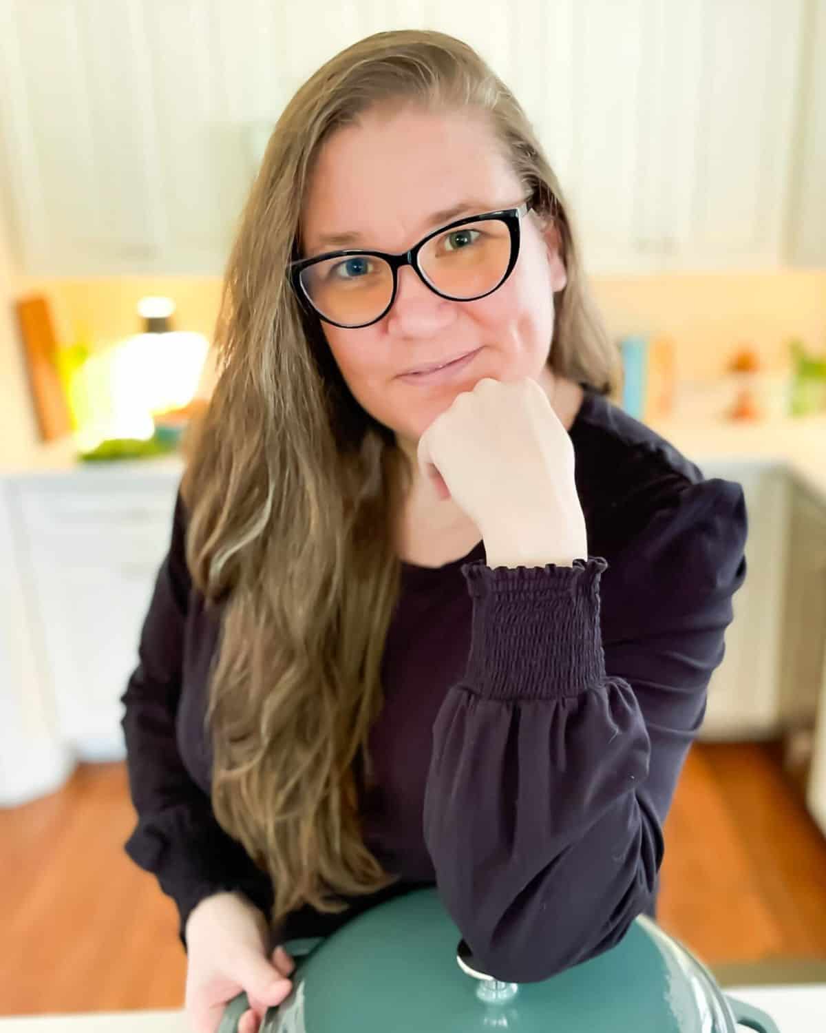 A busy cook with long hair and glasses, wearing a dark top, rests her chin on her hand and stands in the kitchen with one hand on a green pot lid, contemplating her next quick meal.