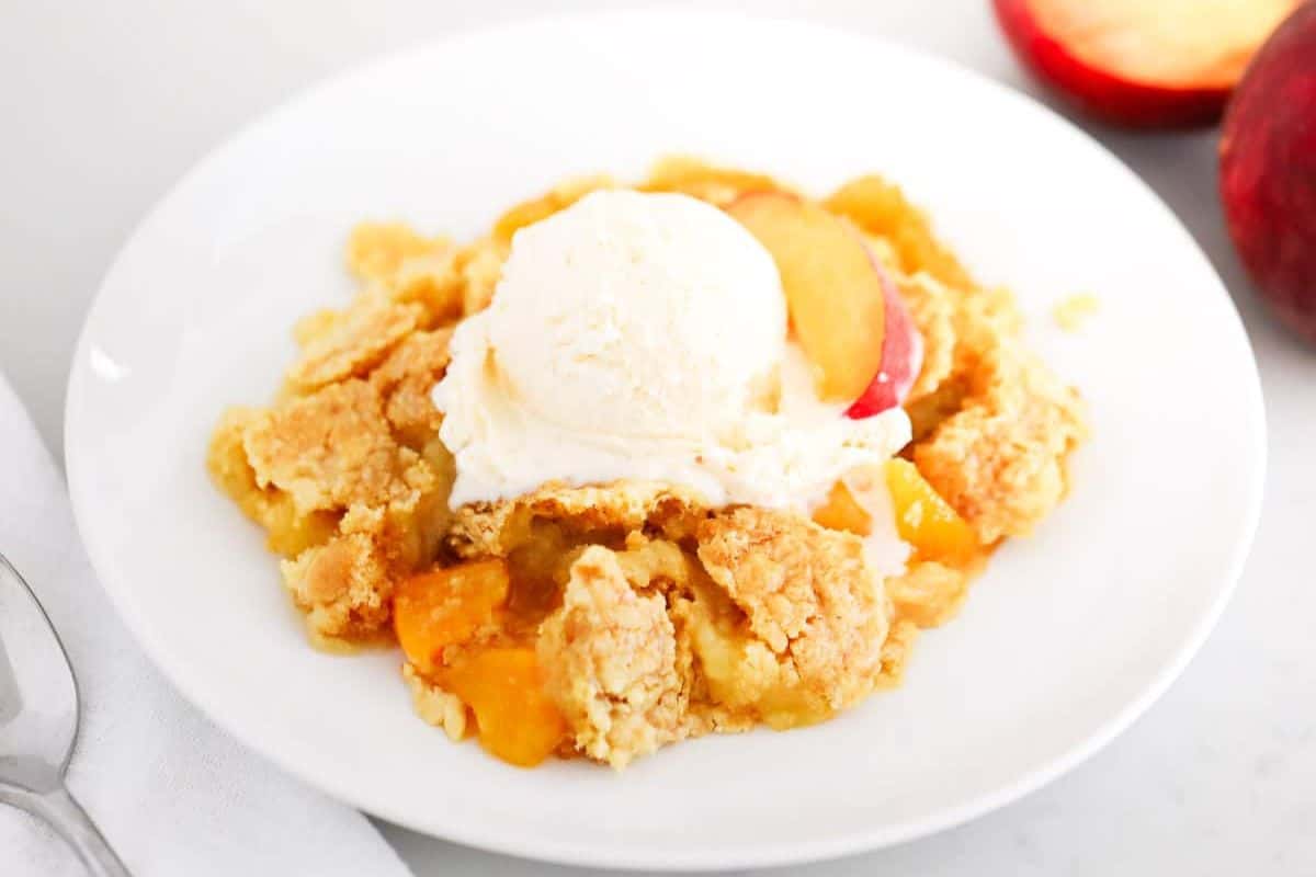 A serving of peach cobbler topped with a scoop of vanilla ice cream on a white plate, with a peach slice and spoon beside it—one of those easy dessert recipes that turns any meal into a wow dessert.