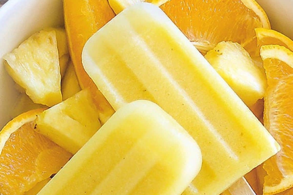 Two yellow popsicles rest on a bed of fresh orange and pineapple slices in a white bowl, creating a vibrant treat to beat the heat with these refreshing popsicles.