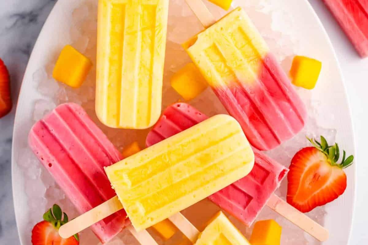 A plate of yellow, pink, and yellow-pink popsicles on ice, surrounded by strawberry halves and mango cubes—refreshing treats to beat the heat and inspire your next popsicle recipes.
