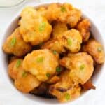 A bowl of golden-brown vegetables garnished with chopped green onions, with a bowl of creamy dipping sauce in the background.
