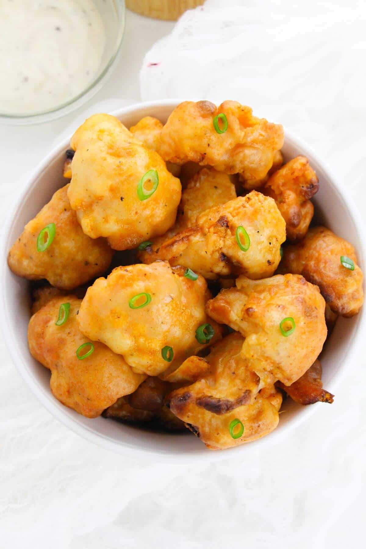 A bowl of golden-brown vegetables garnished with chopped green onions, with a bowl of creamy dipping sauce in the background.