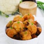 A white bowl filled with Air Fryer Cauliflower Wings garnished with chopped green onions. A head of cauliflower and seasonings are in the background.