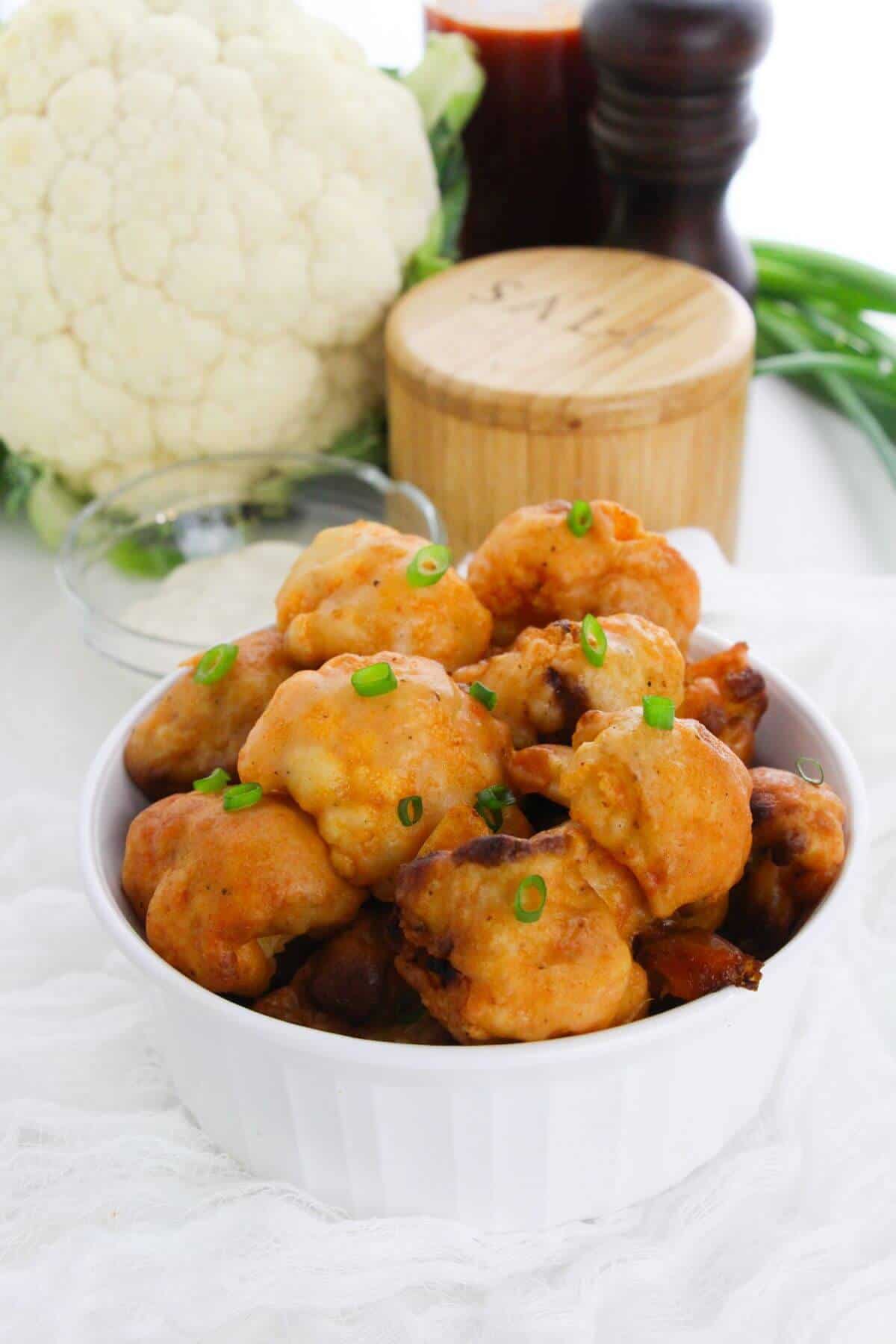 A white bowl filled with Air Fryer Cauliflower Wings garnished with chopped green onions. A head of cauliflower and seasonings are in the background.