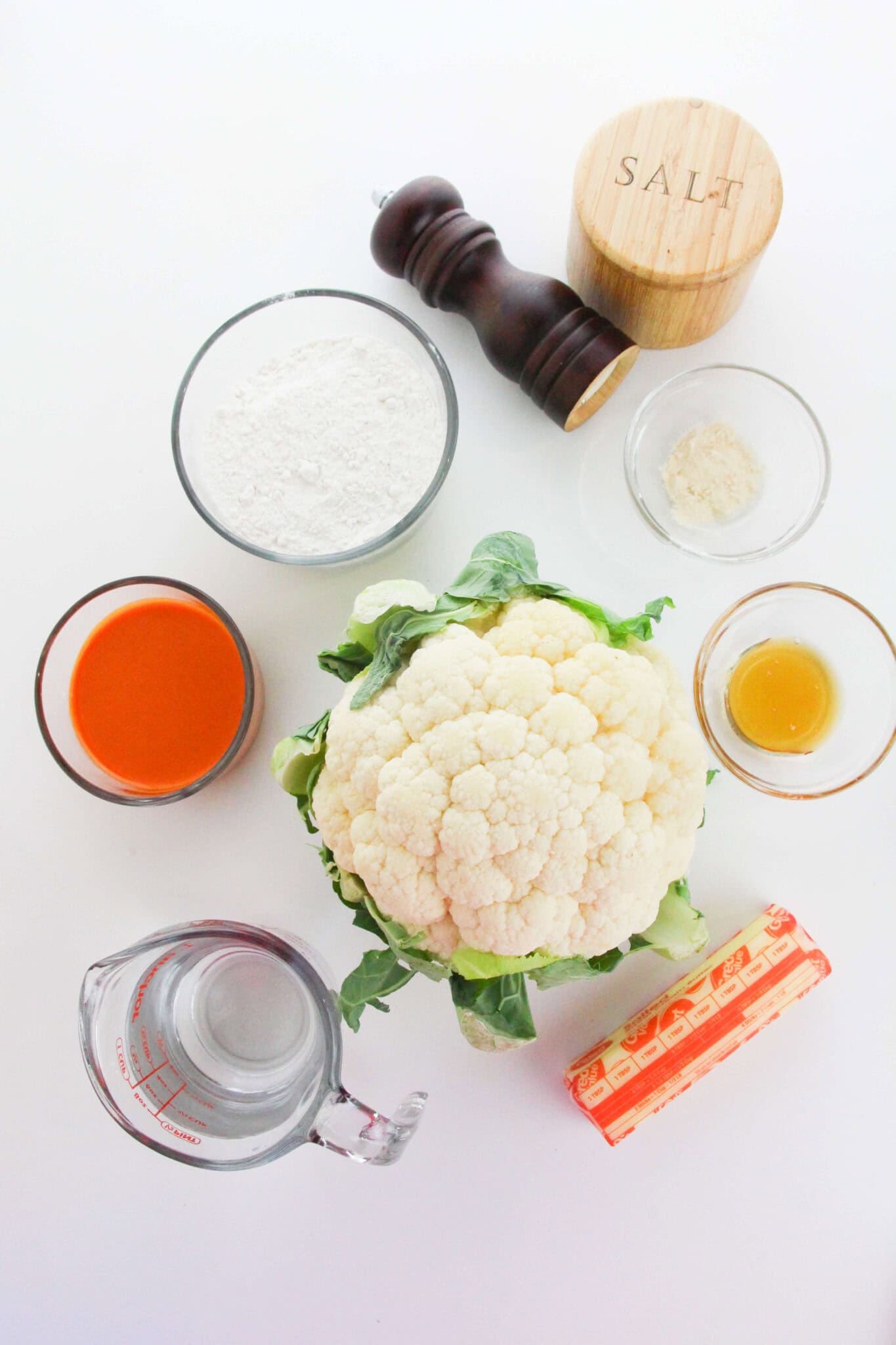 A head of cauliflower surrounded by flour, hot sauce, water, melted butter, salt, black pepper, and garlic powder—perfect ingredients for making crispy Air Fryer Cauliflower Wings—are all arranged on a white surface.