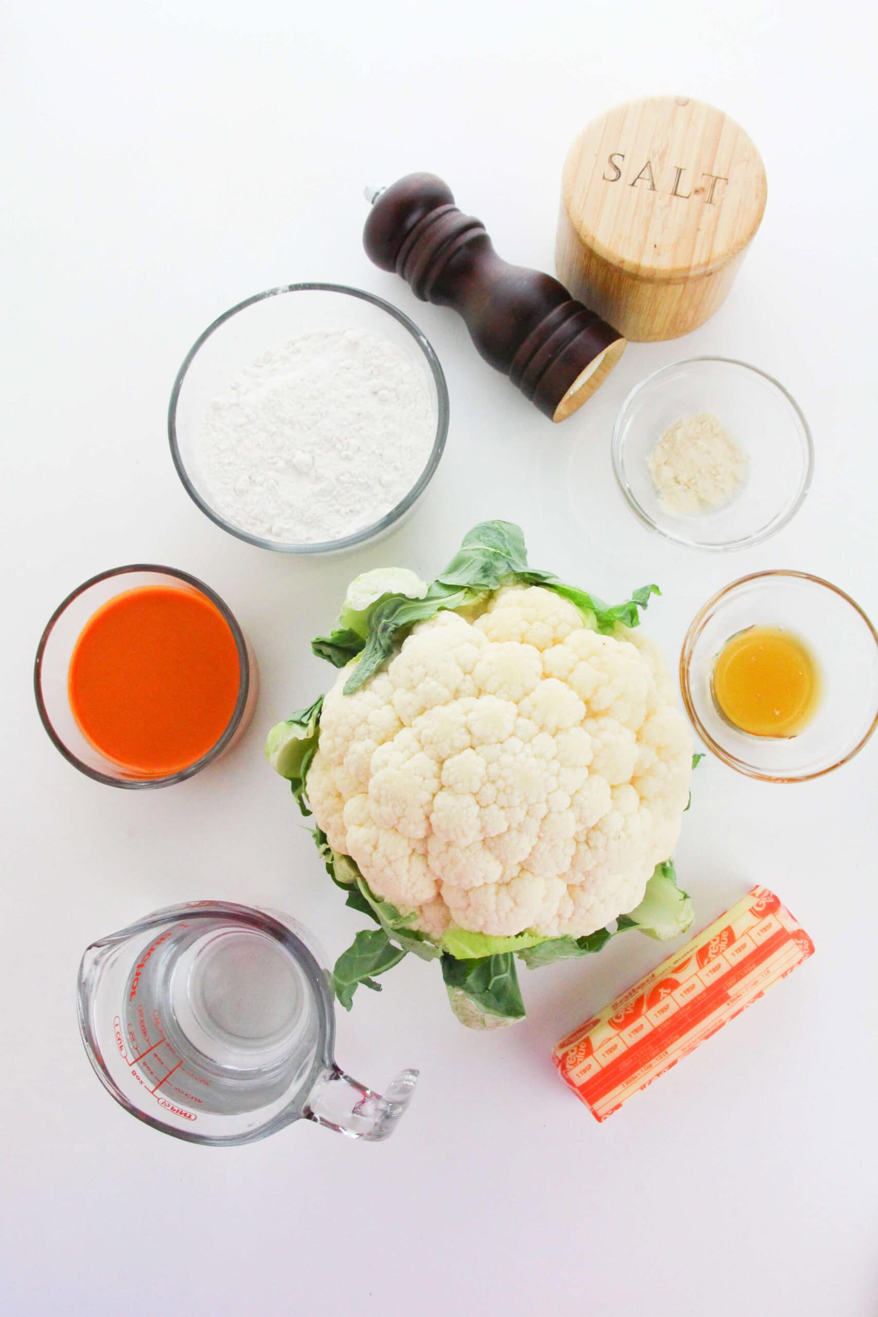 A head of cauliflower surrounded by flour, hot sauce, water, melted butter, salt, black pepper, and garlic powder—perfect ingredients for making crispy Air Fryer Cauliflower Wings—are all arranged on a white surface.