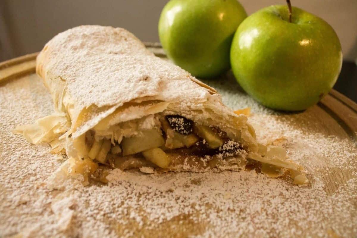A sliced apple strudel, one of the German favorites, is covered in powdered sugar on a tray, with two whole green apples in the background.