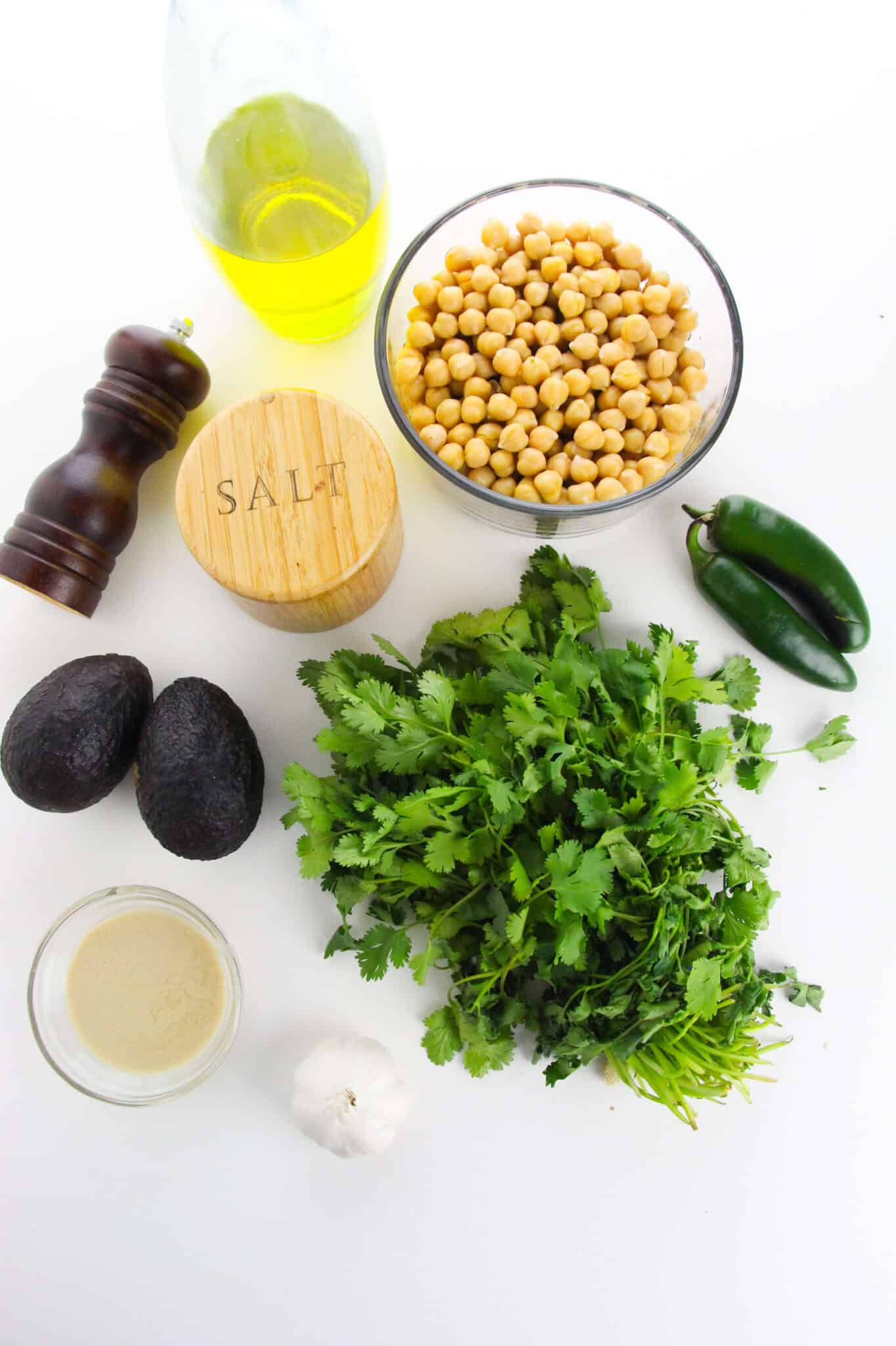 Ingredients for Avocado Hummus, including chickpeas, avocados, cilantro, jalapeños, garlic, olive oil, tahini, salt, and black pepper are arranged on a white surface.
