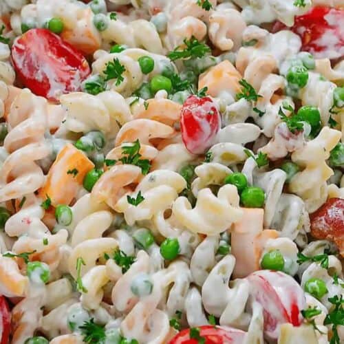 Close-up of creamy pasta salad with rotini, cherry tomatoes, green peas, cheddar cheese cubes, and chopped parsley—a perfect addition to your Memorial Day or Labor Day patriotic party foods lineup.