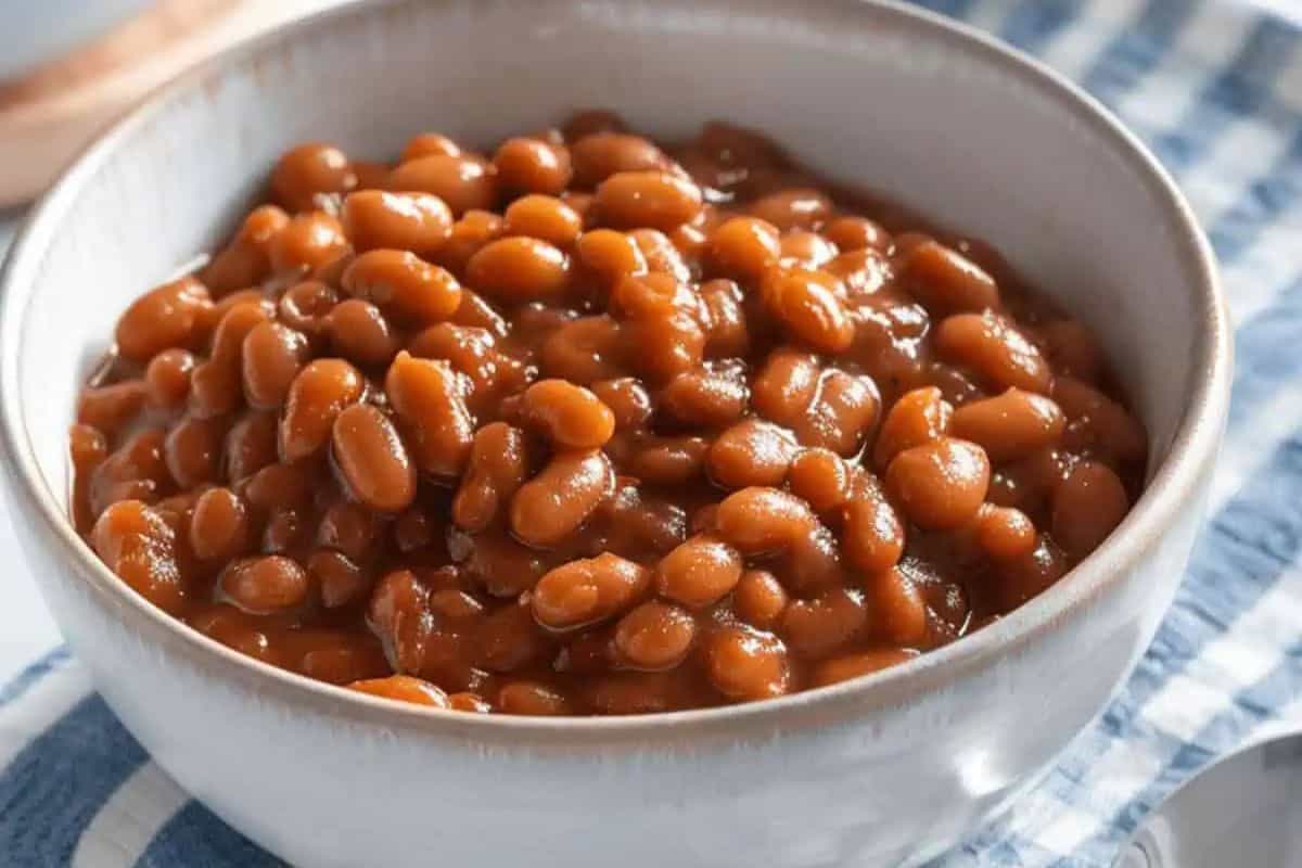 A bowl filled with baked beans in sauce is placed on a blue and white checkered cloth, perfect for classic American recipes or your next BBQ gathering.