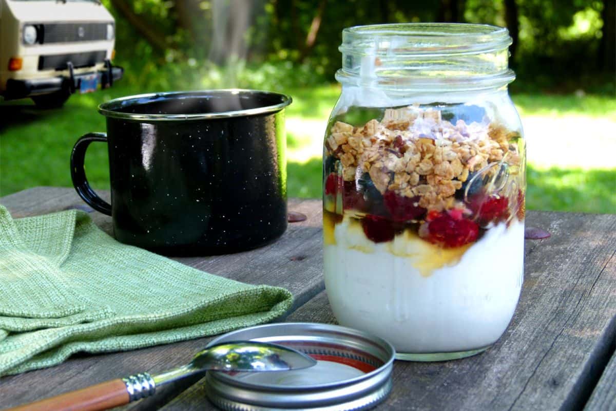 A mason jar filled with flavor-packed yogurt, fruit and granola sits on a picnic table next to a black mug, a green cloth, a spoon and a jar lid, with a van and greenery in the background.