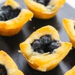 Mini Brie Bites: Small golden pastry cups filled with blueberry filling, arranged on a dark surface.