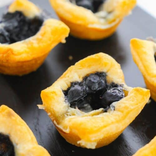 Mini Brie Bites: Small golden pastry cups filled with blueberry filling, arranged on a dark surface.