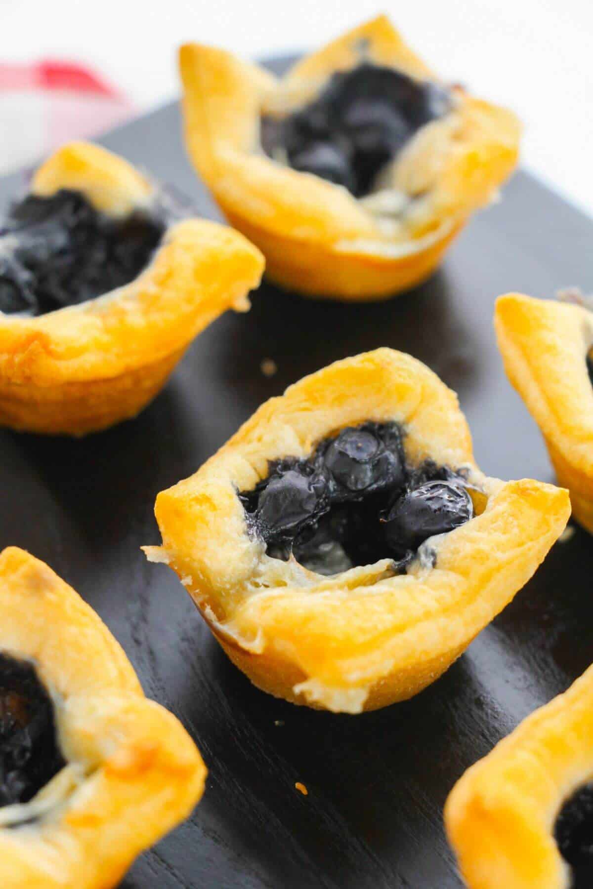 Mini Brie Bites: Small golden pastry cups filled with blueberry filling, arranged on a dark surface.