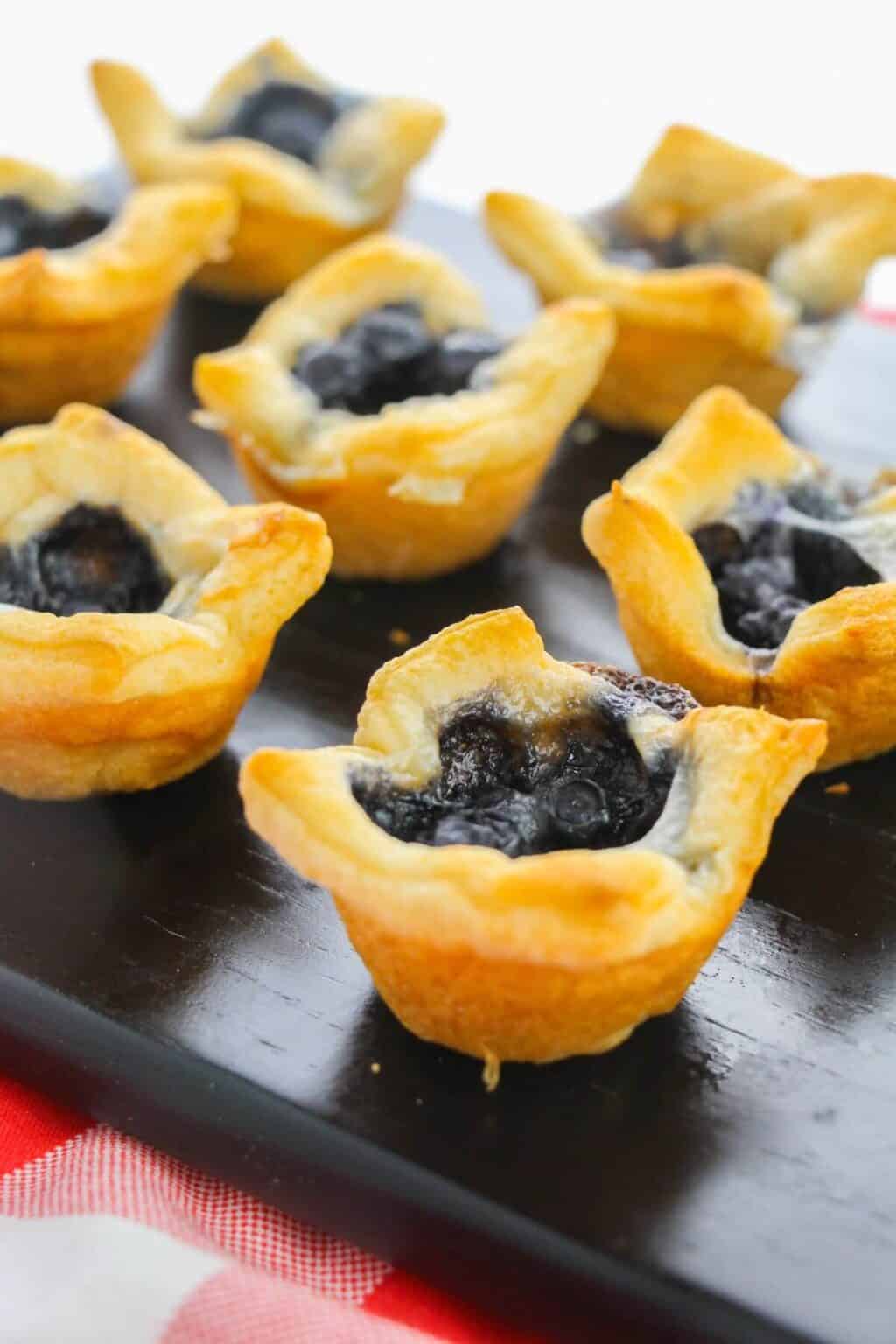 Tarts with golden crusts are arranged on a black serving tray atop a red and white checkered surface, alongside the appetizer.