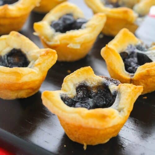 Tarts with golden crusts are arranged on a black serving tray atop a red and white checkered surface, alongside the appetizer.