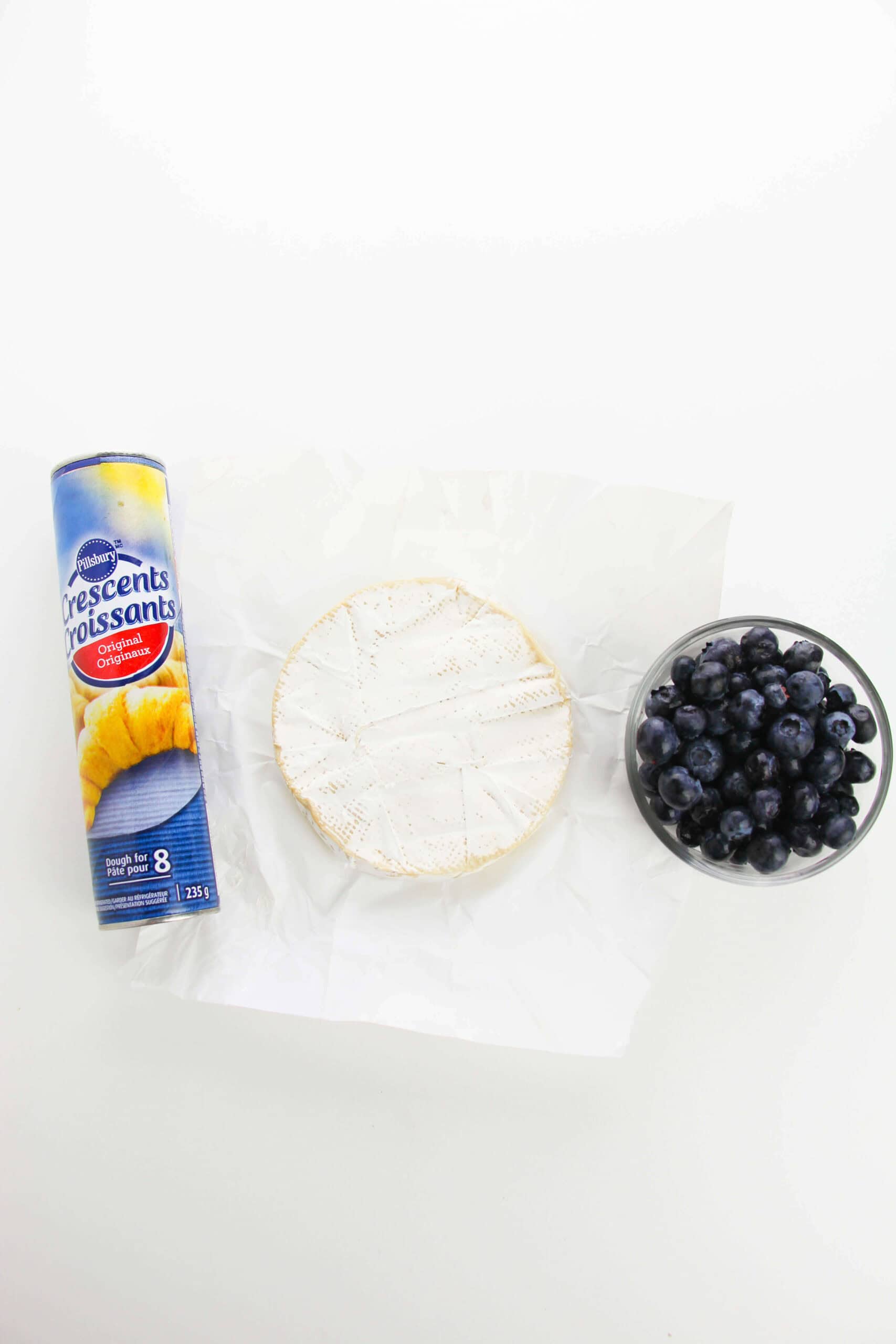 A can of crescent rolls, a wheel of brie cheese on parchment paper, and a glass bowl of blueberries sit on a white surface—perfect for making Mini Brie Bites.