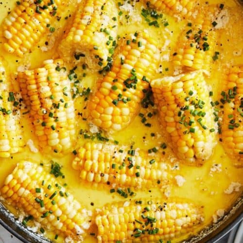 Corn on the cob simmering in a creamy, buttery sauce, topped with chopped chives, served in a large round skillet—perfect for summer recipes.