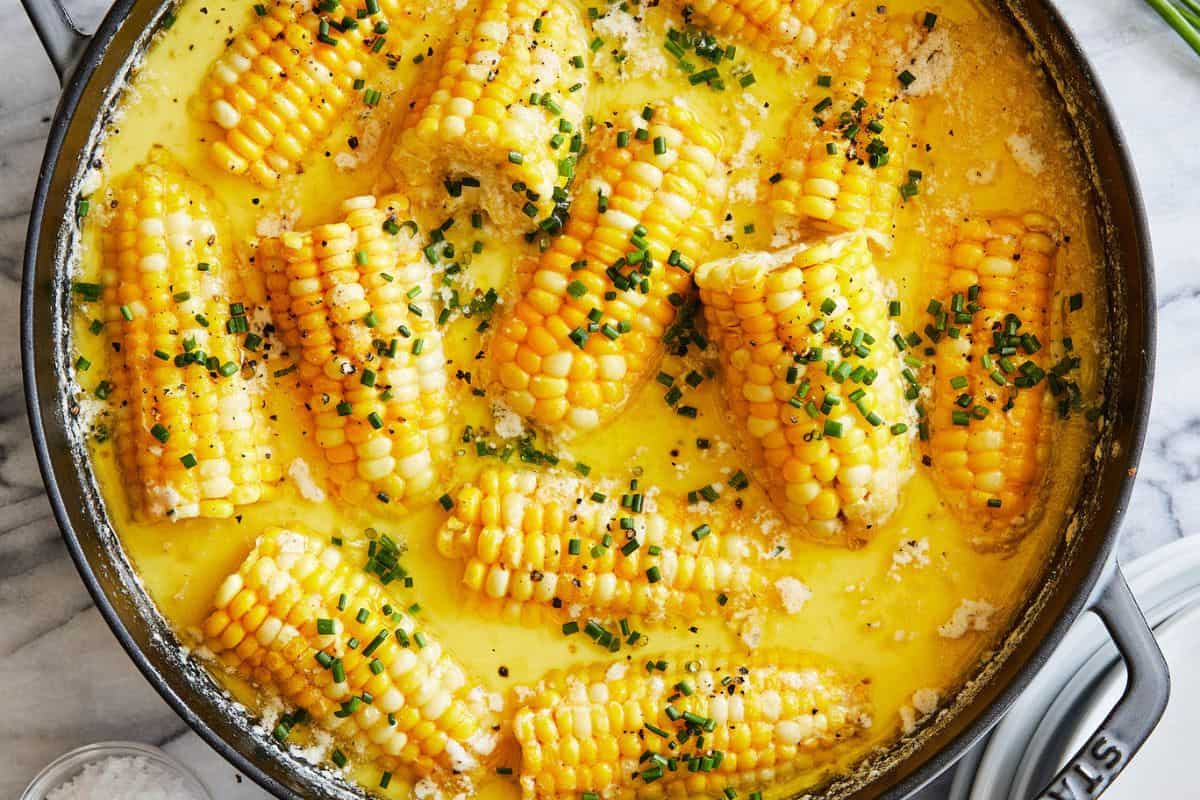 Corn on the cob simmering in a creamy, buttery sauce, topped with chopped chives, served in a large round skillet—perfect for summer recipes.