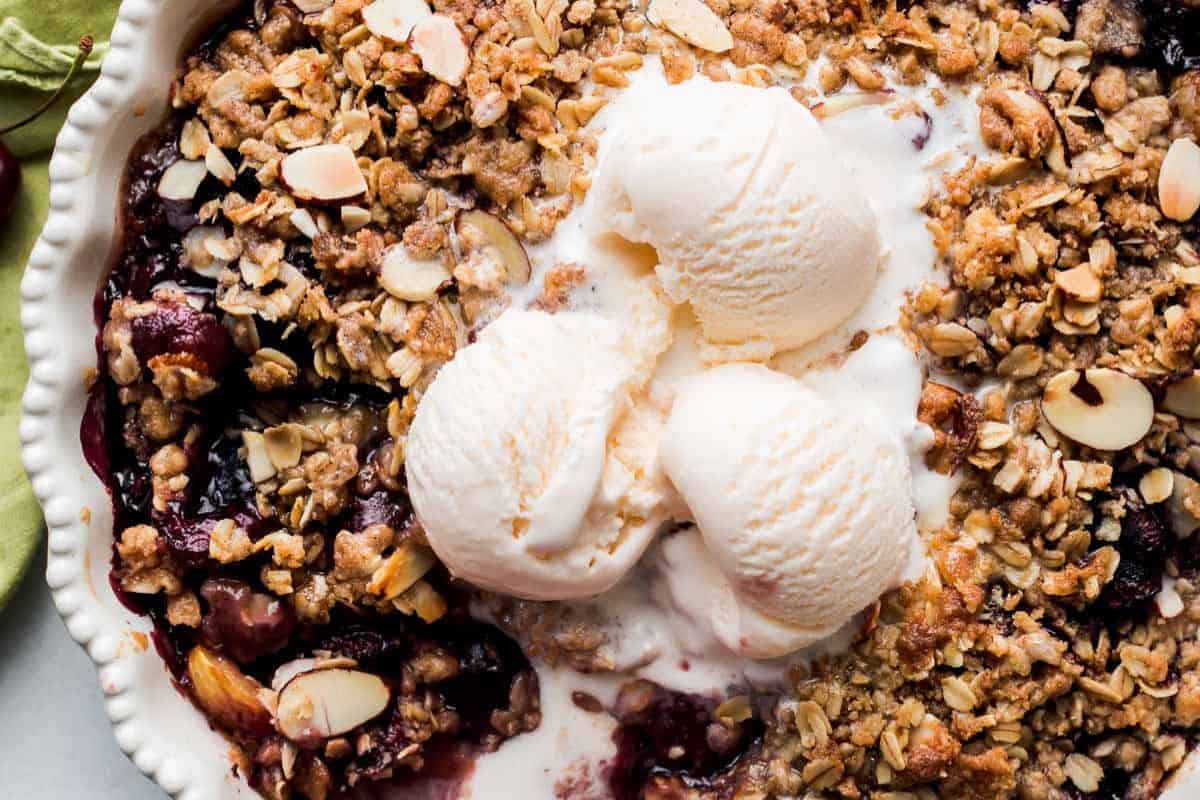 A baked fruit crumble featuring fresh cherry, topped with oats, sliced almonds, and three scoops of vanilla ice cream in a white dish.