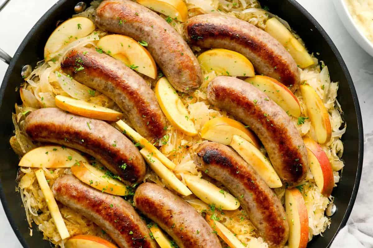 Cooked sausages arranged on a bed of sauerkraut with sliced apples in a skillet, garnished with chopped herbs—a perfect way to enjoy German favorites or bring Oktoberfest flavors home with easy recipes.